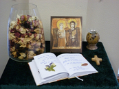 Eine Ablage ist zur Totenerinnerung gestaltet mit einer Vase voller Trockenblumen, einer Ikone, ein Holzkreuz, ein Kerzengefäß und ein Erinnerungsbuch.