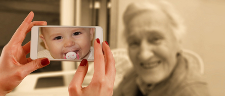 Das Bild zeigt Hände, die ein Smartphone halten. Auf dem Bildschirm des Smartphones ist ein Baby mit Schnuller deutlich zu erkennen. Im unscharfen Hintergrund ist schemenhaft das Gesicht einer älteren Frau zu erkennen, die lächelt. Die Farben des Hinte Das Bild zeigt Hände, die ein Smartphone halten. Auf dem Bildschirm des Smartphones ist ein Baby mit Schnuller deutlich zu erkennen. Im unscharfen Hintergrund ist schemenhaft das Gesicht einer älteren Frau zu erkennen, die lächelt. Die Farben des Hintergrunds sind sepiafarben.