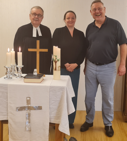 Der Pfarrer und sein Team stehen hinter dem vorbereiteten Altar in einem Wohnraum. Der Pfarrer und sein Team stehen hinter dem vorbereiteten Altar in einem Wohnraum.