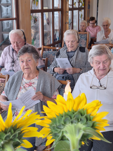 Ein Bild zeigt eine Gruppe älterer Menschen, die an einer Veranstaltung teilnehmen und Texte von Blättern lesen. Einige tragen Kopfhörer und sind in Stühle versenkt. Im Vordergrund sind Sonnenblumen und eine Kerzenlampe zu sehen, die eine fröhliche und ruhige Atmosphäre schaffen.